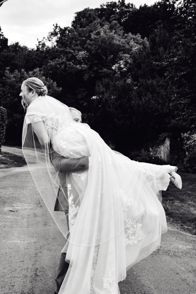 Braut trägt ihren Bräutigam auf einer Landstraße, umgeben von Bäumen.