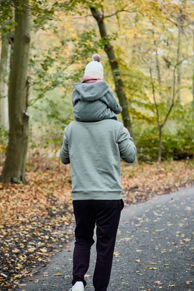 Eine Person trägt ein Kind auf den Schultern und geht einen Waldweg entlang.