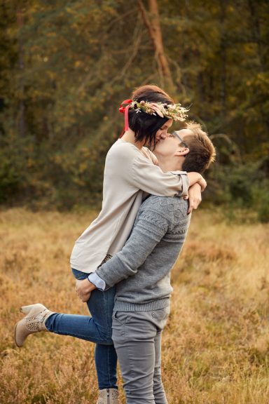 Paar umarmt sich in freier Natur, Frau in blauer Jeans und grauem Pullover, mit Blumenkranz.