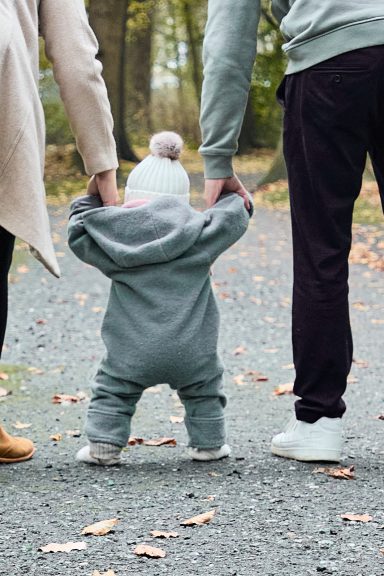 Kleines Kind in grauem Anzug hält sich an den Händen der Eltern auf einem schmalen Weg.