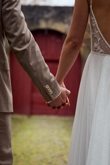 Hochzeitskleid und Anzughände halten sich fest. Hintergrund mit roter Wand.