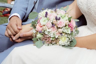 Hände eines Paares halten sich, während eine blumige Brautsträuße sichtbar ist.