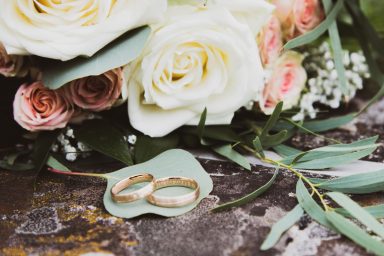 Hochzeitsringe auf einem grünen Blatt, umgeben von einer Blumenarrangement in sanften Farben.