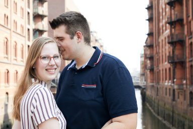Ein Paar posiert lächelnd vor einem Kanal und roten Backsteingebäuden.