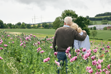 Älteres Paar steht in einem Blumenfeld und blickt auf eine grüne Landschaft.